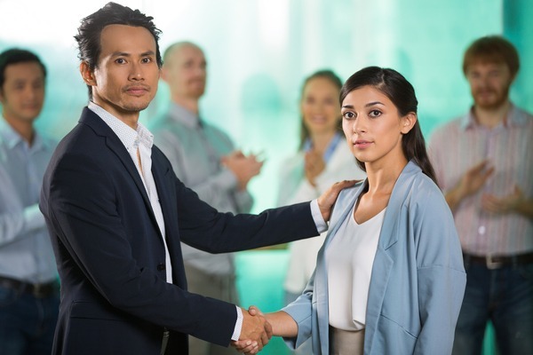 A imagem contém um advogado do Direito Trabalhista dando um aperto de mão e apoiando o ombro de sua cliente. Ao fundo os colegas de trabalho batem palmas.